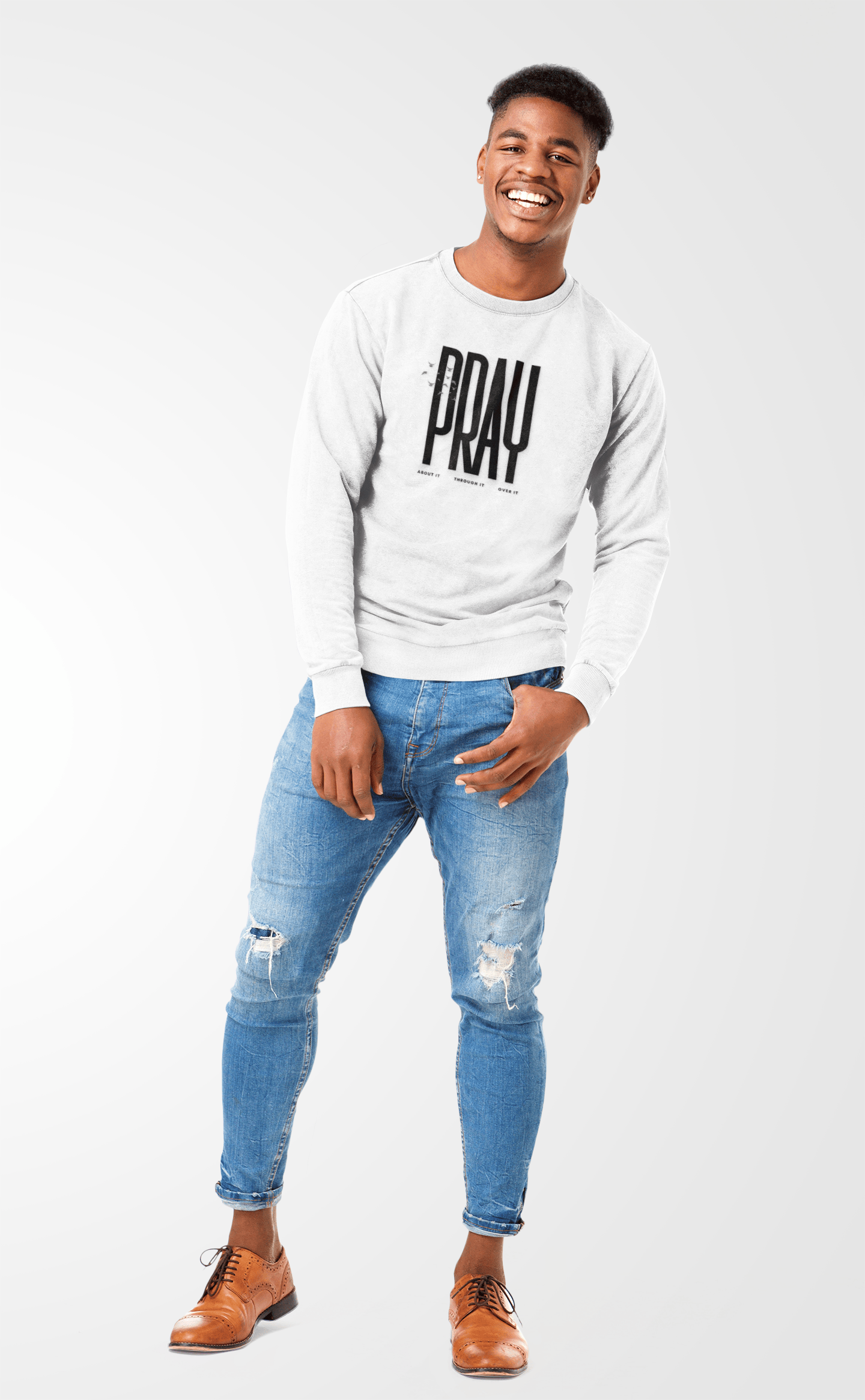 Man wearing a white sweatshirt with 'PRAY' printed on it, blue jeans, and brown shoes against a white background.